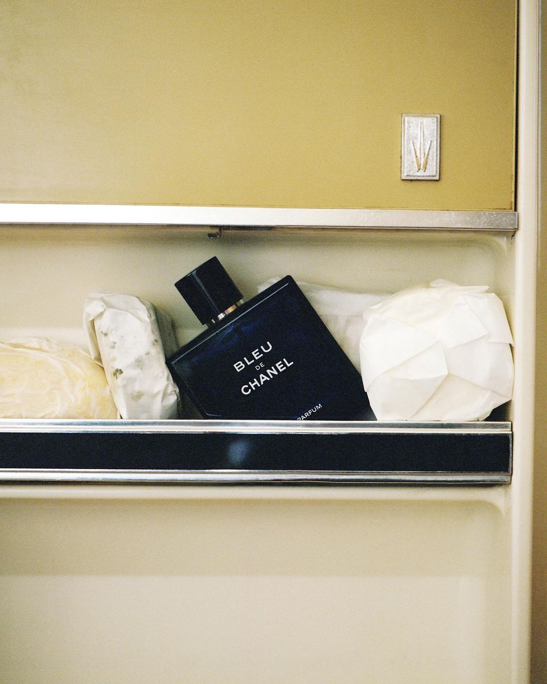Still life of Bleu de Chanel perfume bottle resting on a shelf alongside wrapped food items, illuminated by soft, diffused lighting.
