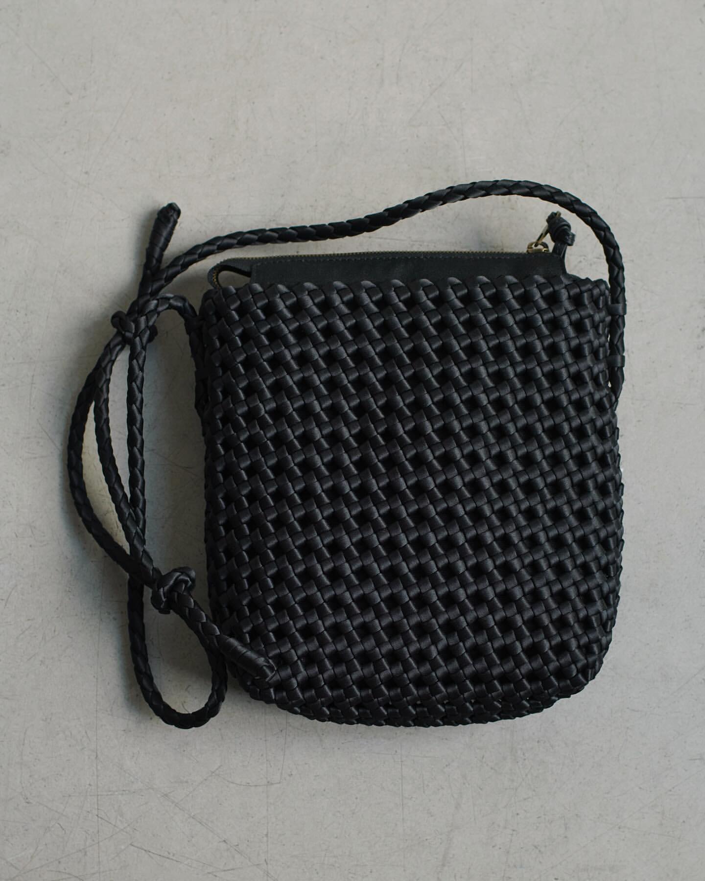 Black woven crossbody bag with a braided shoulder strap on a neutral surface, product photography showing texture and design.