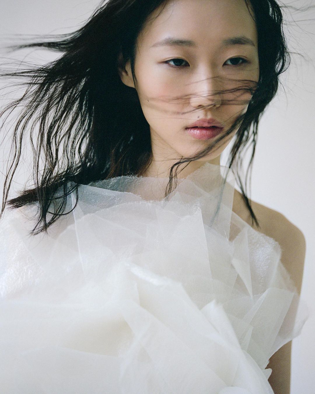 Portrait of an Asian woman with flowing dark hair wearing a white layered tulle garment against a light background in a photo studio.