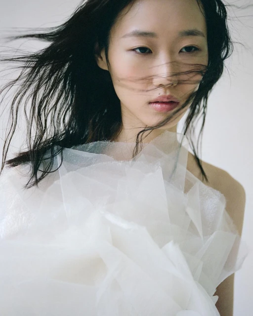 Portrait of an Asian woman with flowing dark hair wearing a white layered tulle garment against a light background in a photo studio.