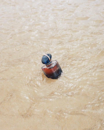 A perfume bottle resting in shallow water over light sandy ripples, photographed by Aude Le Barbey in Paris.