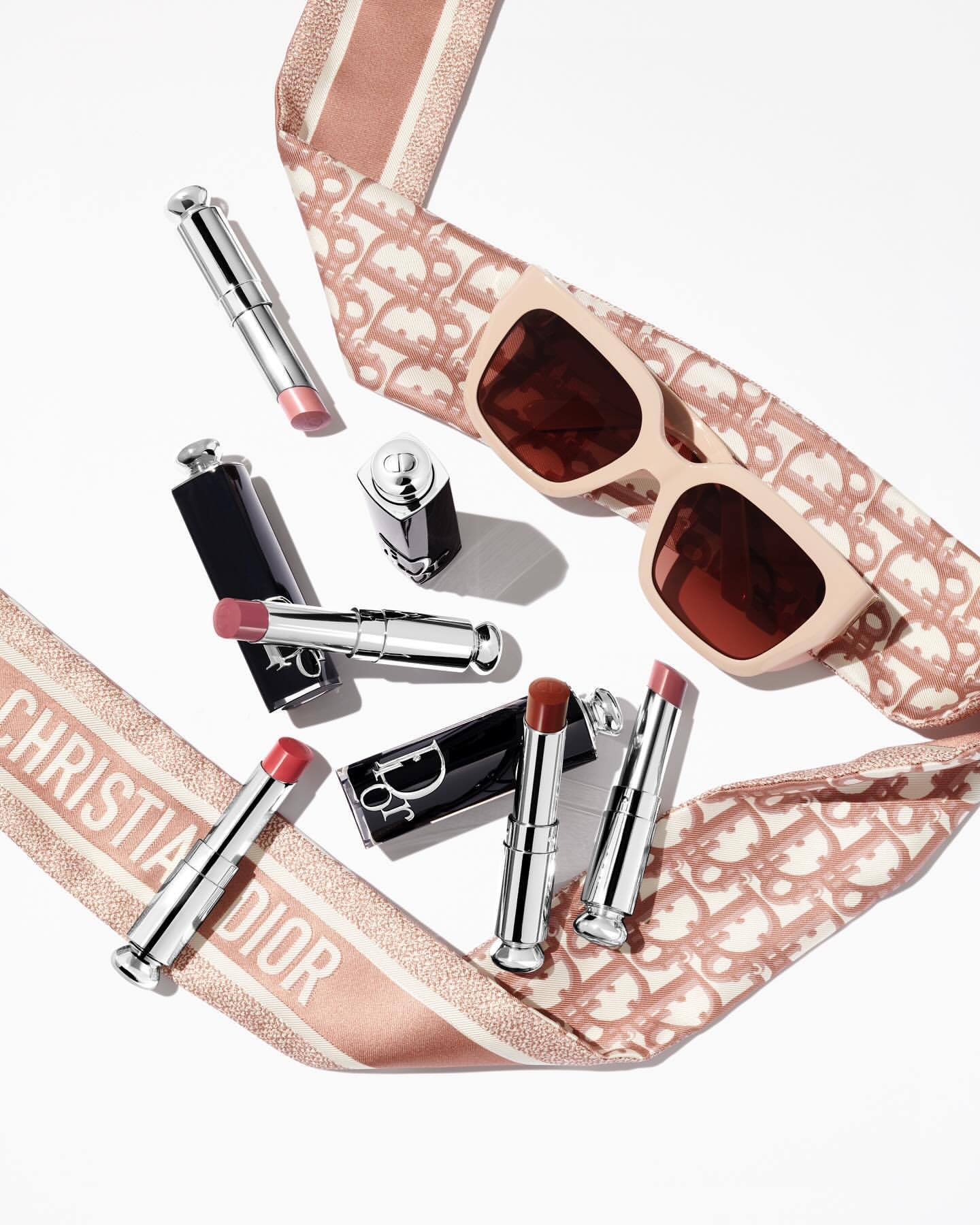 Flat lay arrangement of Dior lipsticks and sunglasses framed by a patterned pink ribbon on white background
