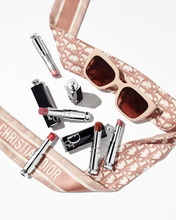 Flat lay arrangement of Dior lipsticks and sunglasses framed by a patterned pink ribbon on white background
