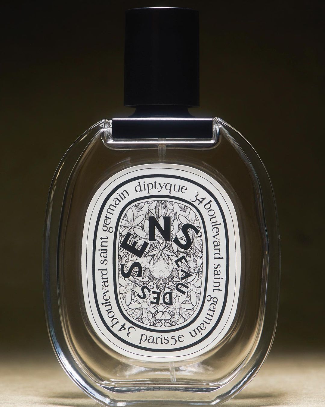 Diptyque 'Eau des Sens' perfume bottle with oval clear glass body, black cap, and white label, isolated against a blurred dark backdrop.