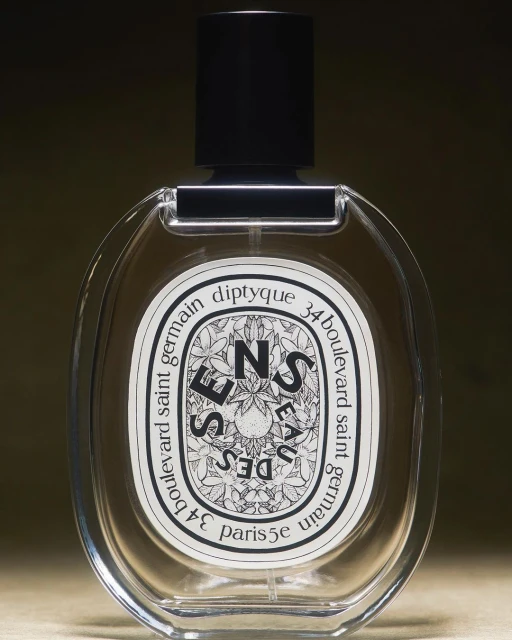 Diptyque 'Eau des Sens' perfume bottle with oval clear glass body, black cap, and white label, isolated against a blurred dark backdrop.