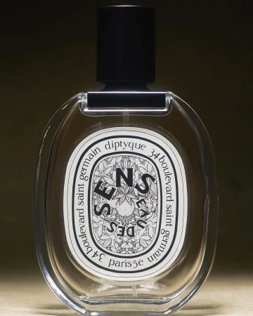 Diptyque 'Eau des Sens' perfume bottle with oval clear glass body, black cap, and white label, isolated against a blurred dark backdrop.