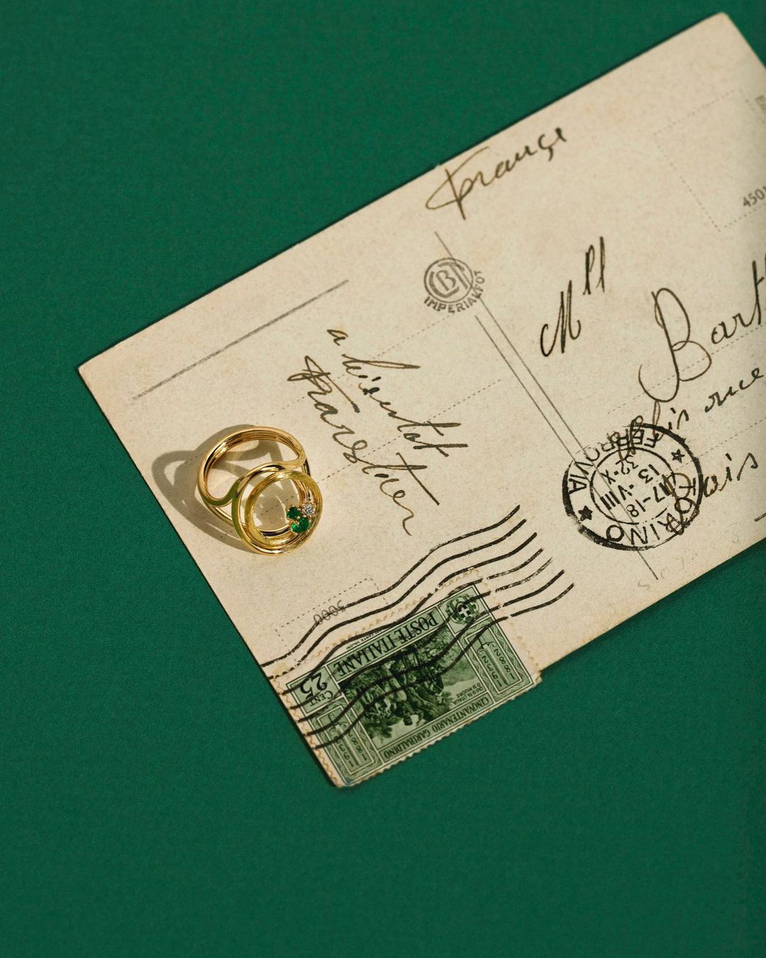 Gold ring with emeralds and diamonds on a vintage postcard with handwritten text, presented on a green background. Jewelry close-up.