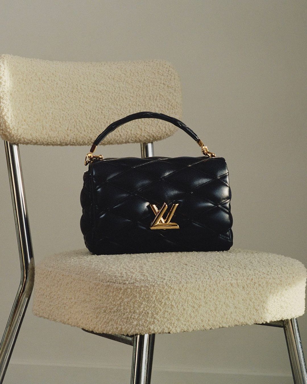 Quilted black leather handbag with gold hardware and logo on a cream-colored sherpa chair against a light neutral backdrop.