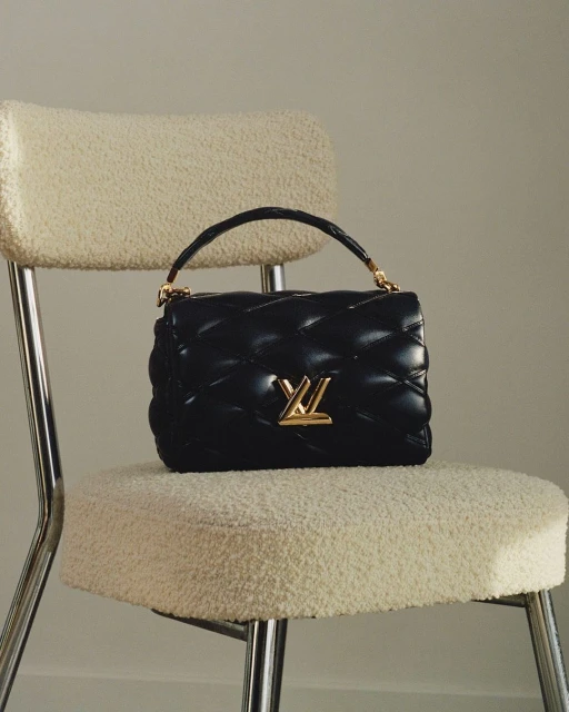 Quilted black leather handbag with gold hardware and logo on a cream-colored sherpa chair against a light neutral backdrop.