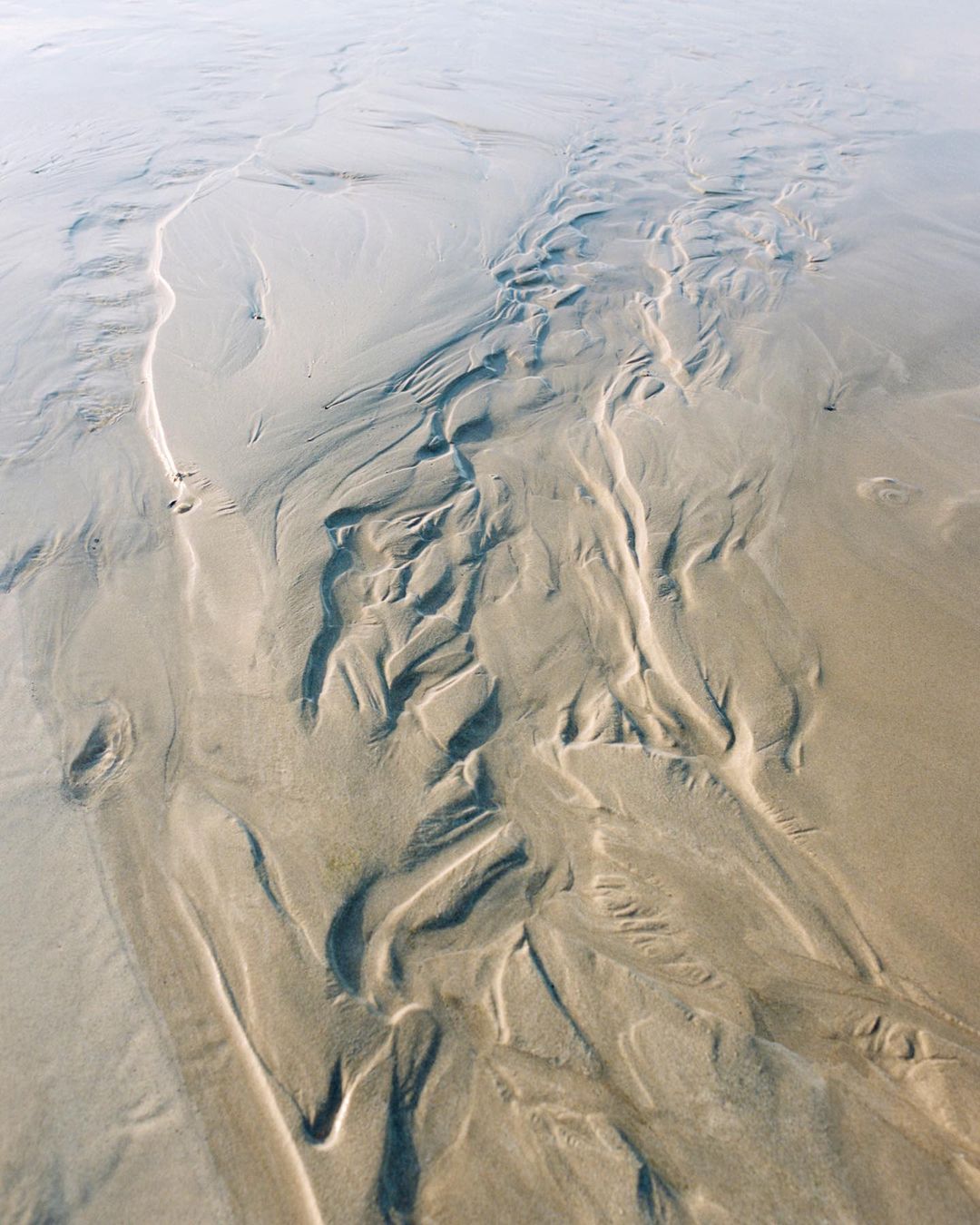 Aerial view of wet sandy beach with abstract ripple patterns and tidal streams during low tide, with light reflecting on the surface.