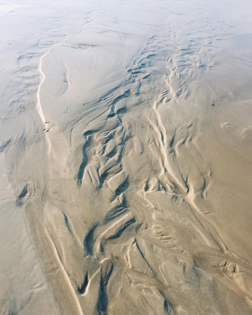 Aerial view of wet sandy beach with abstract ripple patterns and tidal streams during low tide, with light reflecting on the surface.