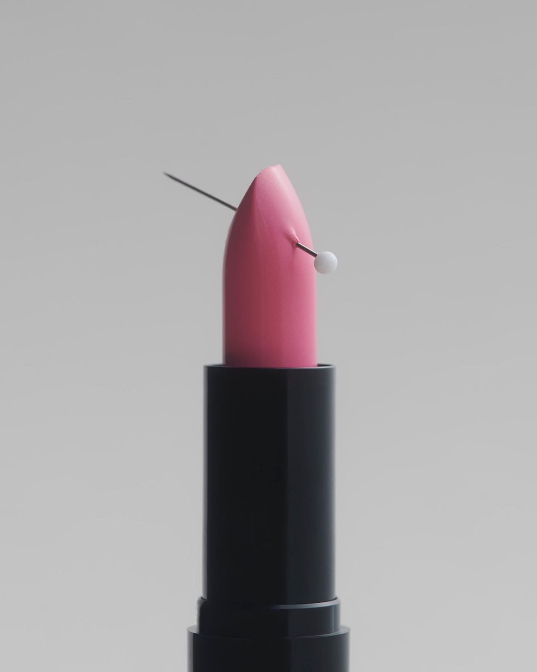 Pink lipstick in a black tube pierced by a white-headed pin against a neutral grey seamless background