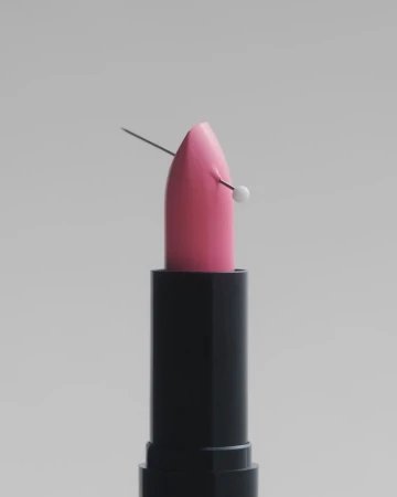 Pink lipstick in a black tube pierced by a white-headed pin against a neutral grey seamless background