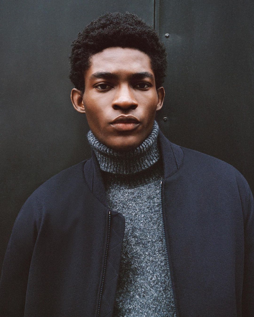 Close-up portrait of a young, Black man wearing a grey turtleneck sweater and a dark navy jacket, against a dark grey background.