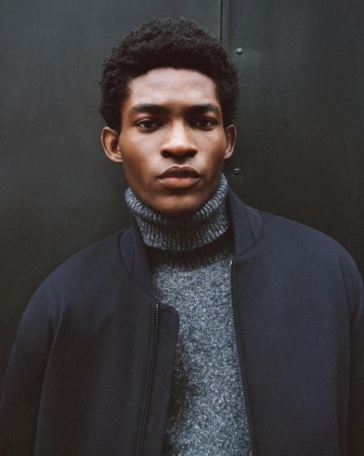 Close-up portrait of a young, Black man wearing a grey turtleneck sweater and a dark navy jacket, against a dark grey background.