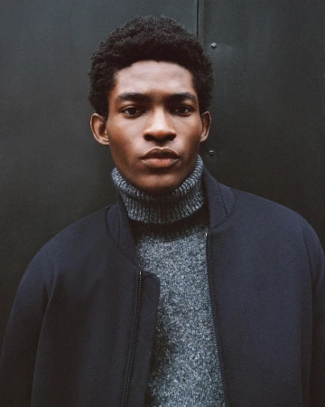 Close-up portrait of a young, Black man wearing a grey turtleneck sweater and a dark navy jacket, against a dark grey background.
