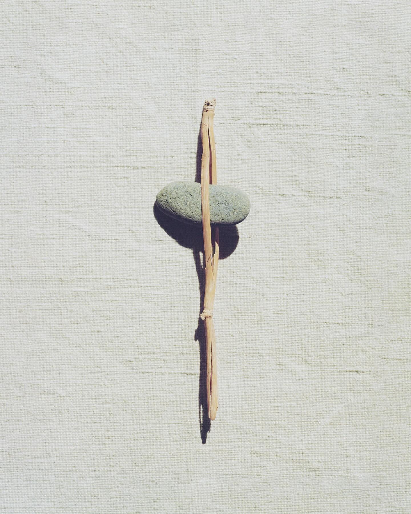 Still life shot of a pale green smooth stone balanced vertically with a broken twig on a white textured surface, minimalist composition.