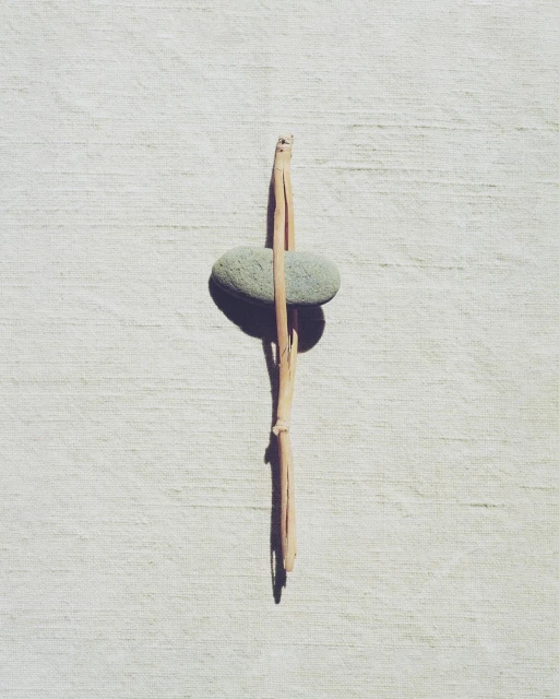 Still life shot of a pale green smooth stone balanced vertically with a broken twig on a white textured surface, minimalist composition.