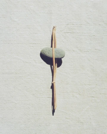 Still life shot of a pale green smooth stone balanced vertically with a broken twig on a white textured surface, minimalist composition.