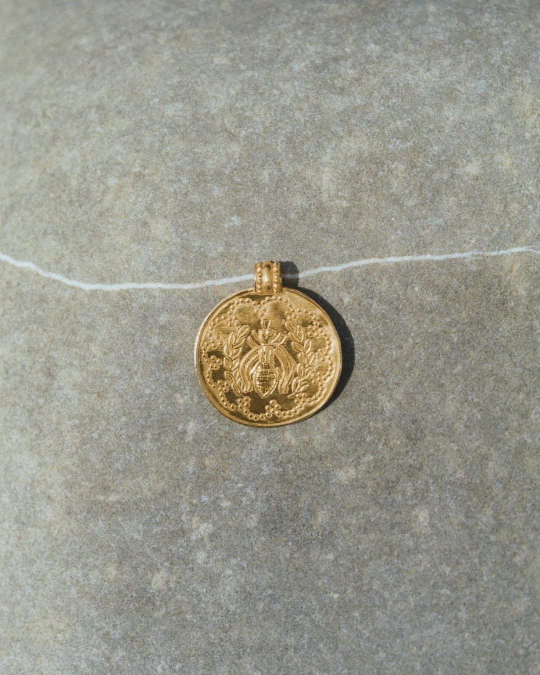 Gold coin pendant featuring bee design on textured gray stone background with thin white string. Jewellery product photography.