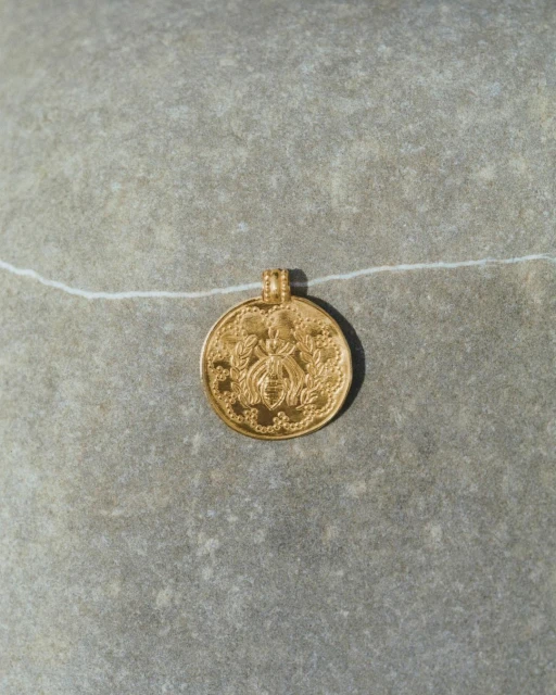 Gold coin pendant featuring bee design on textured gray stone background with thin white string. Jewellery product photography.