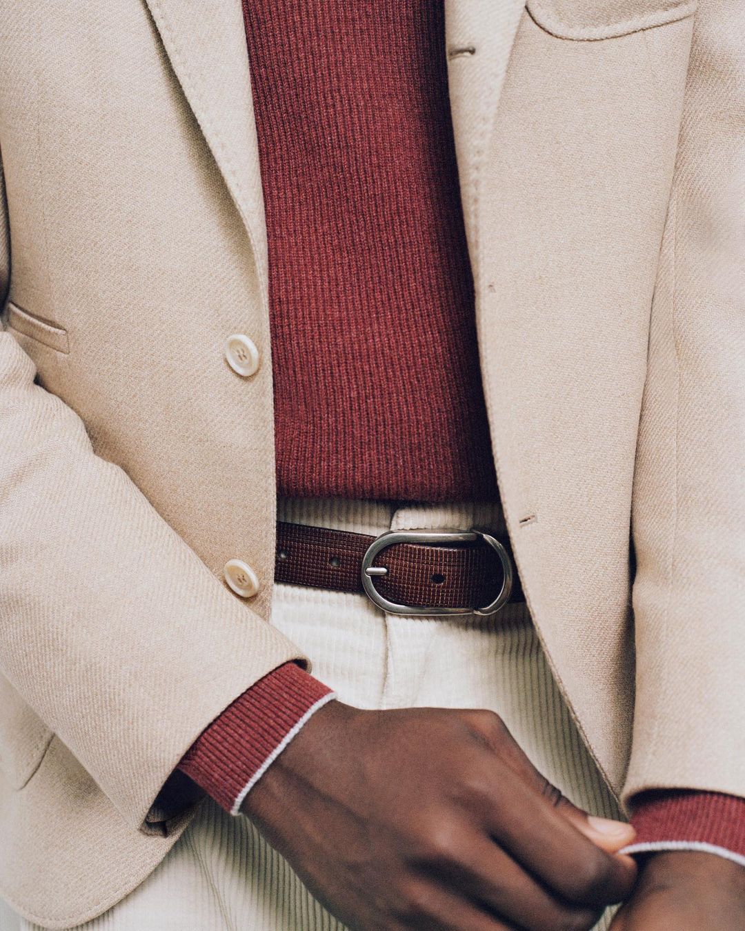 Close-up studio shot of an adult wearing a beige blazer, burgundy sweater, brown belt, and off-white corduroy pants.