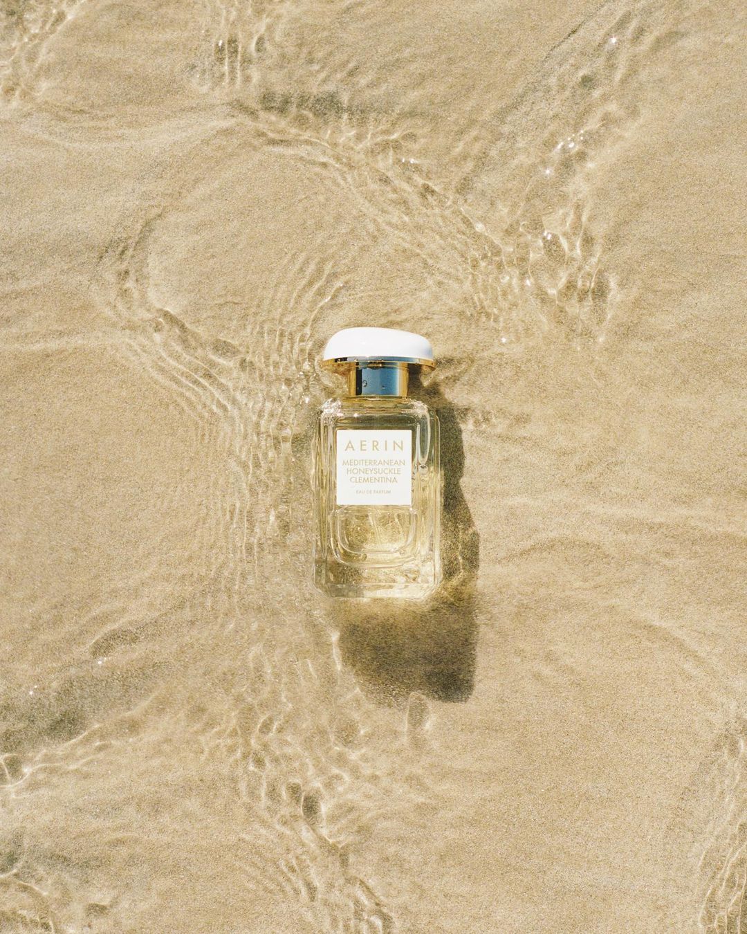 A bottle of AERIN Mediterranean Honeysuckle Clementina perfume sits on a sandy beach with water washing around it in natural light.