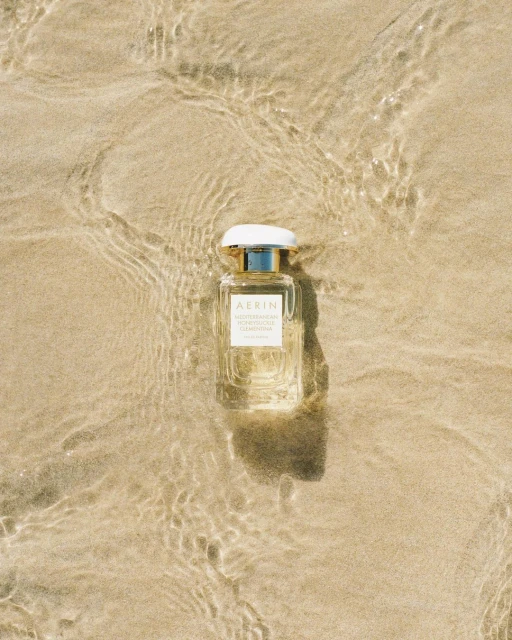 A bottle of AERIN Mediterranean Honeysuckle Clementina perfume sits on a sandy beach with water washing around it in natural light.