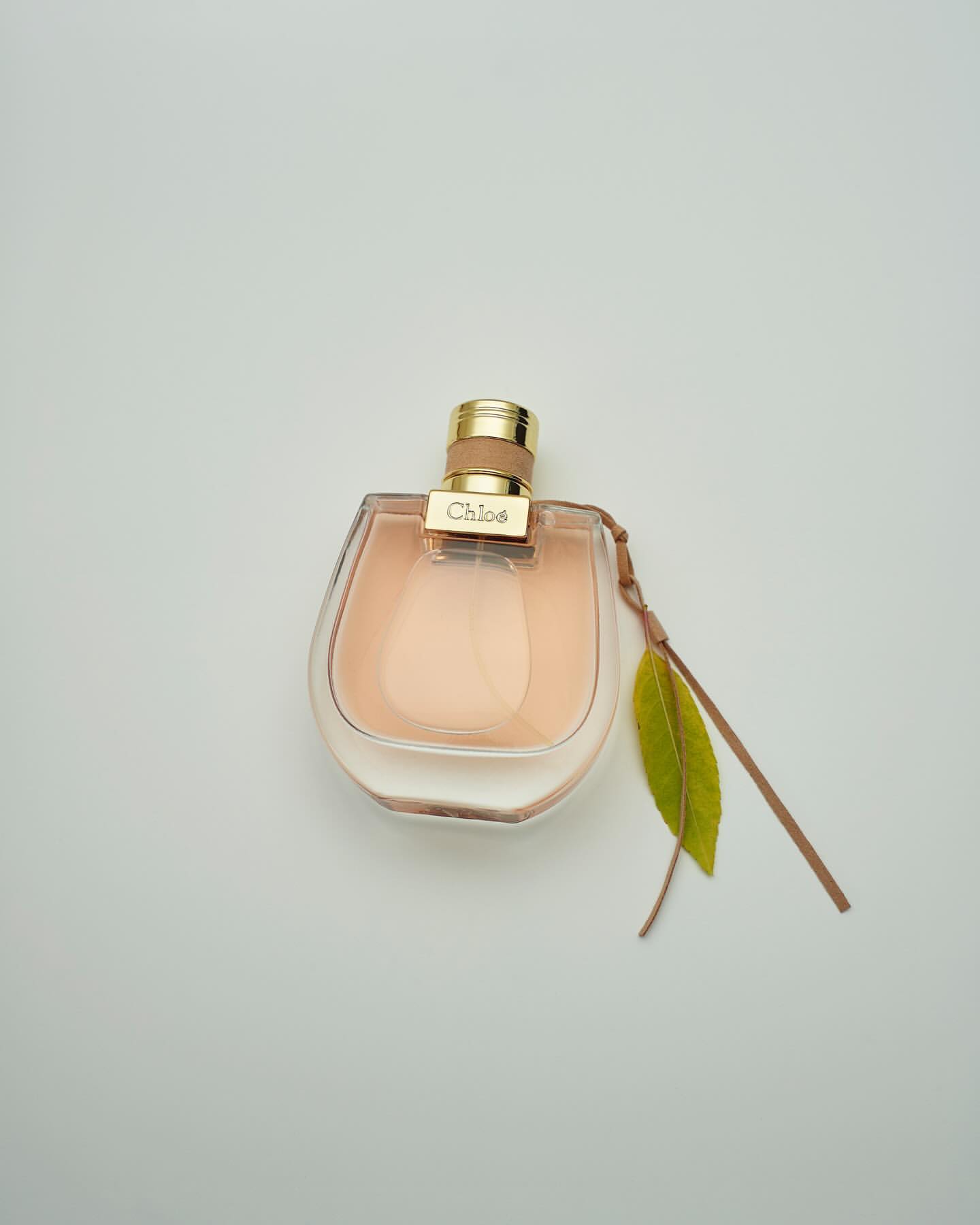 Close-up of a perfume bottle with a gold cap and a green leaf, set against a light background, suggesting a clean and elegant fragrance.