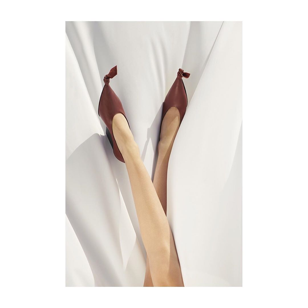 A pair of women's legs extended upward wearing brown ballet flat shoes with tied knots, set against a bright white textile backdrop.
