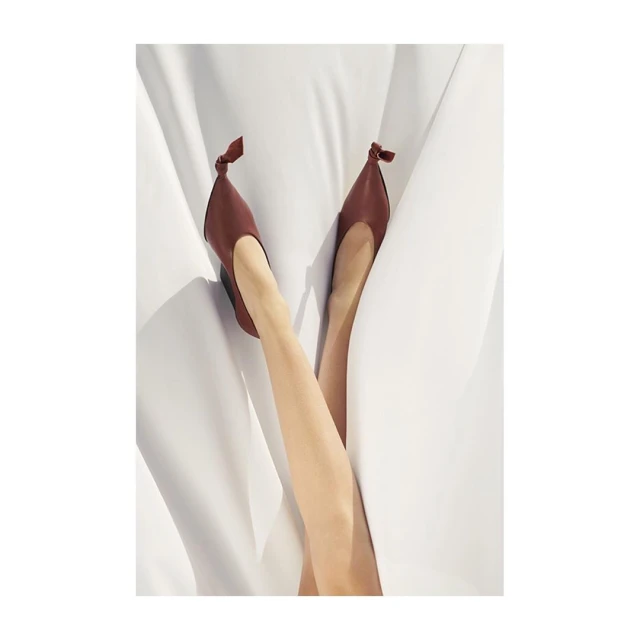 A pair of women's legs extended upward wearing brown ballet flat shoes with tied knots, set against a bright white textile backdrop.