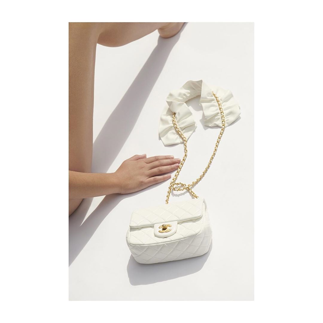 Fashion still life: a person's hand rests near a quilted white handbag with a gold chain on a white surface with a cast shadow.