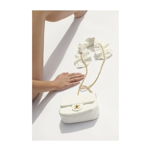Fashion still life: a person's hand rests near a quilted white handbag with a gold chain on a white surface with a cast shadow.