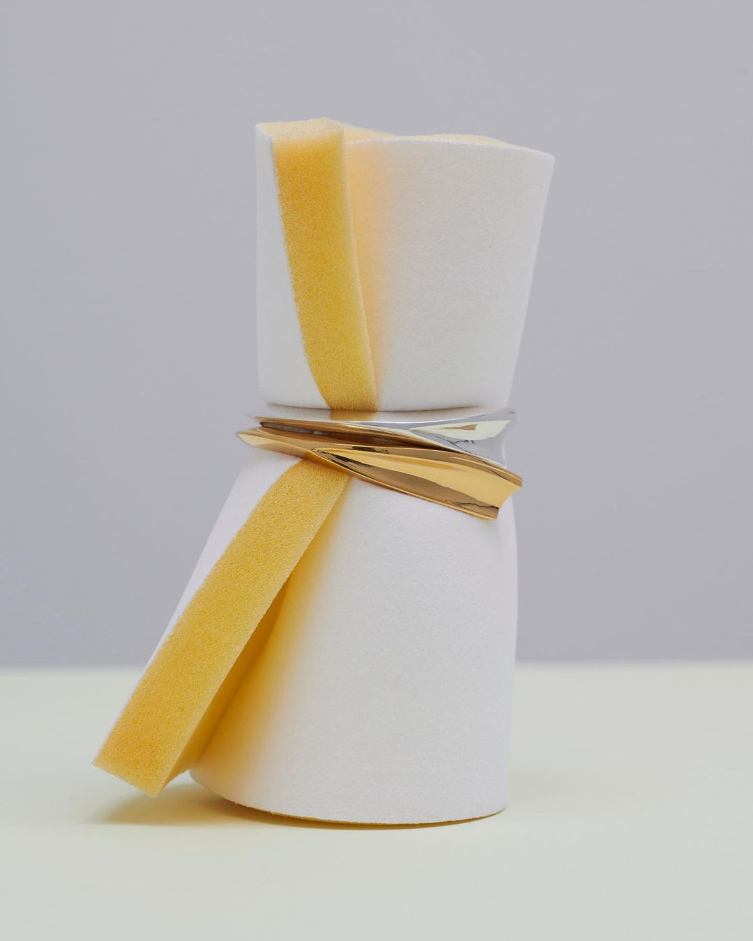 Still life of a white bottle wrapped with a yellow sponge, a silver band, and golden accents, set against a gradient white and grey background.