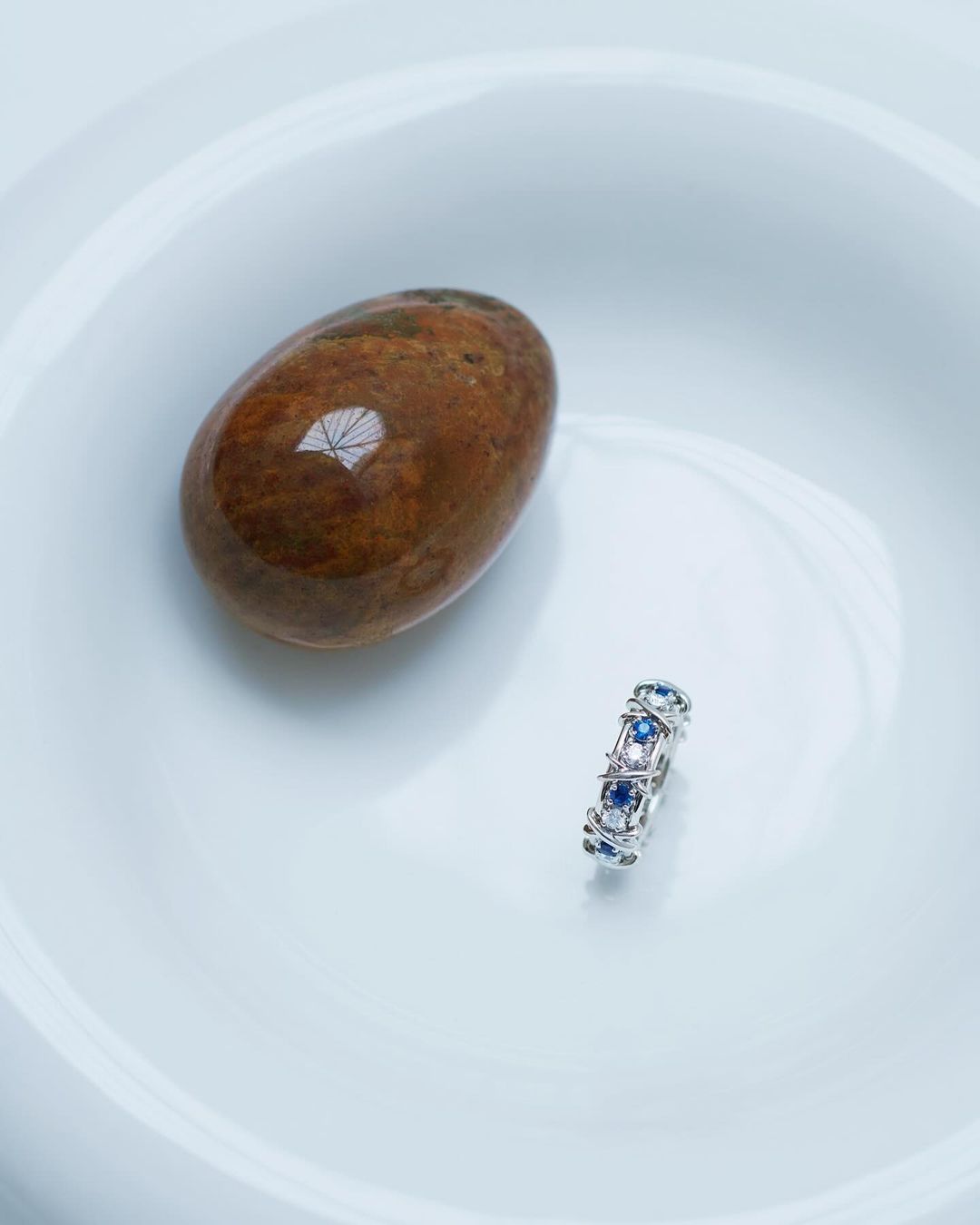 Close-up of an oval brown stone alongside a silver ring with blue and white gemstones, presented on a smooth white surface.