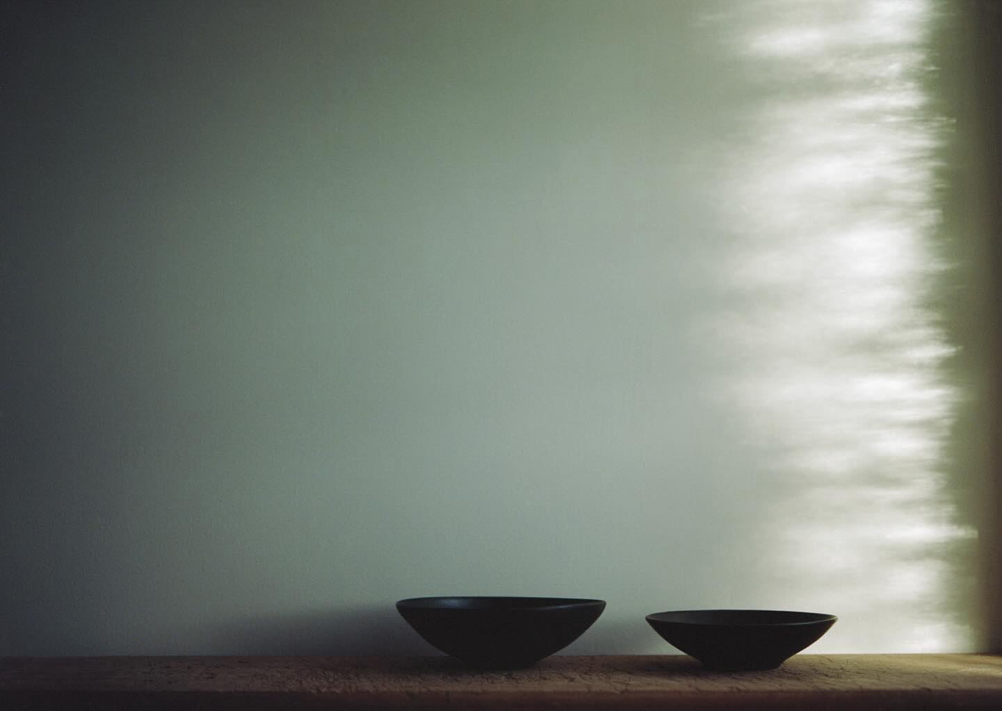 Still life image of two dark ceramic bowls arranged on a wooden surface against a soft grey wall with diffused natural light.