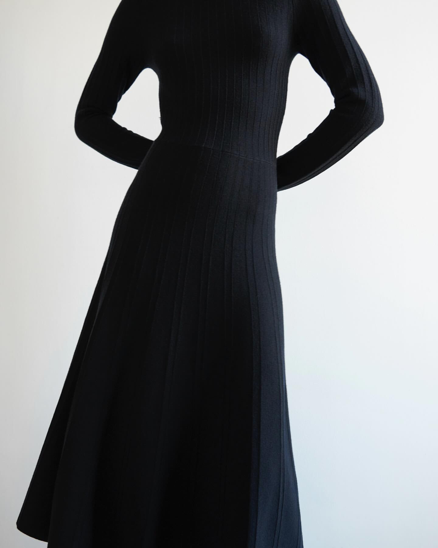 Woman in a long-sleeve black dress with a ribbed texture. The dress flows to mid-calf length against a plain white backdrop.