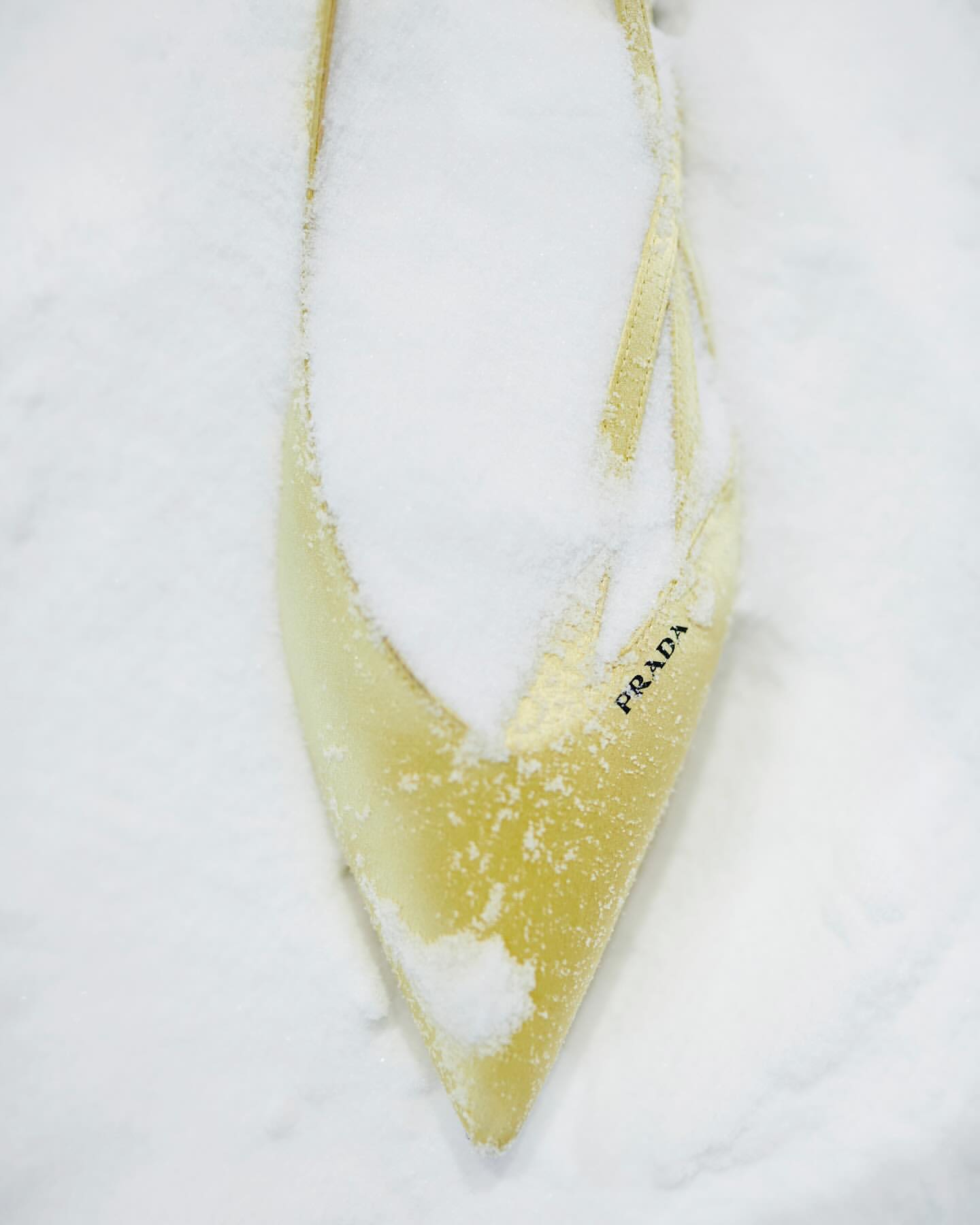 A pointed toe yellow shoe partially covered in fresh snow with visible Prada branding on the side.