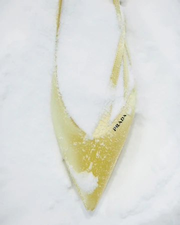 A pointed toe yellow shoe partially covered in fresh snow with visible Prada branding on the side.