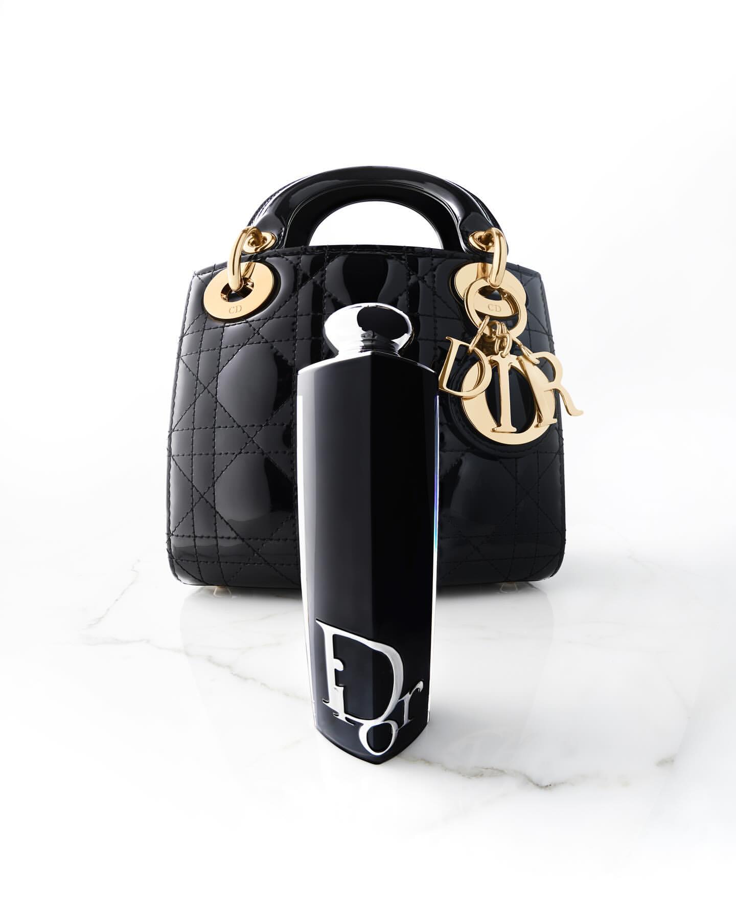 Still life of a black Lady Dior handbag with gold hardware, next to a black Dior perfume bottle with a white logo, on marble.