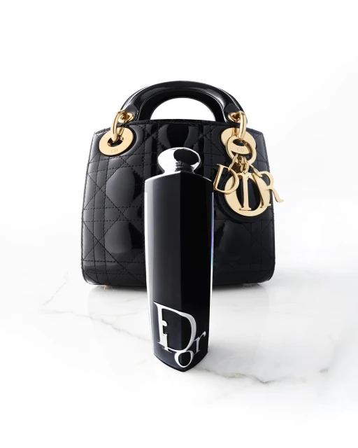 Still life of a black Lady Dior handbag with gold hardware, next to a black Dior perfume bottle with a white logo, on marble.