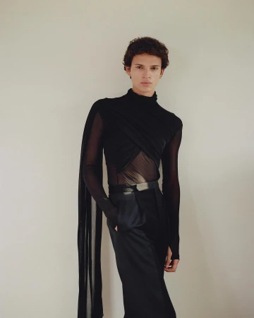 Young male model in a black turtleneck and dark trousers standing against a plain light wall in Paris.