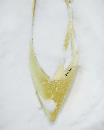 Fashion shot of a light yellow high-heel shoe covered in snow, with a pointed toe and the brand name 'PRADA' visible on its side.