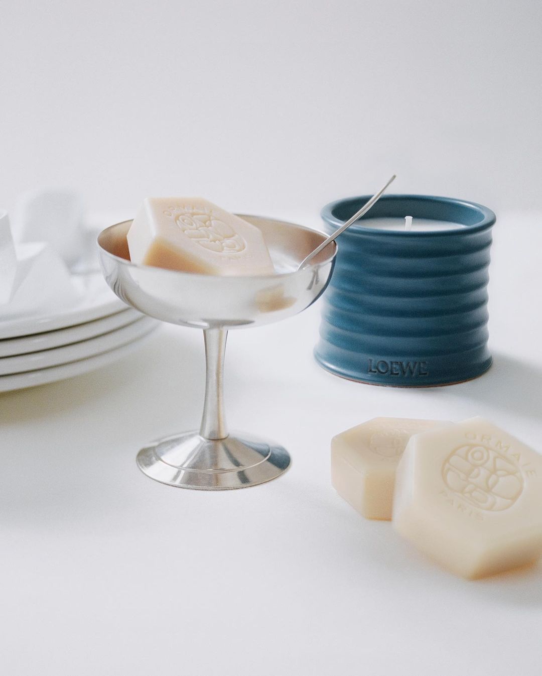 Silver dessert cup with a bar of soap and a spoon, alongside a candle and stacked plates on a white surface in soft lighting.