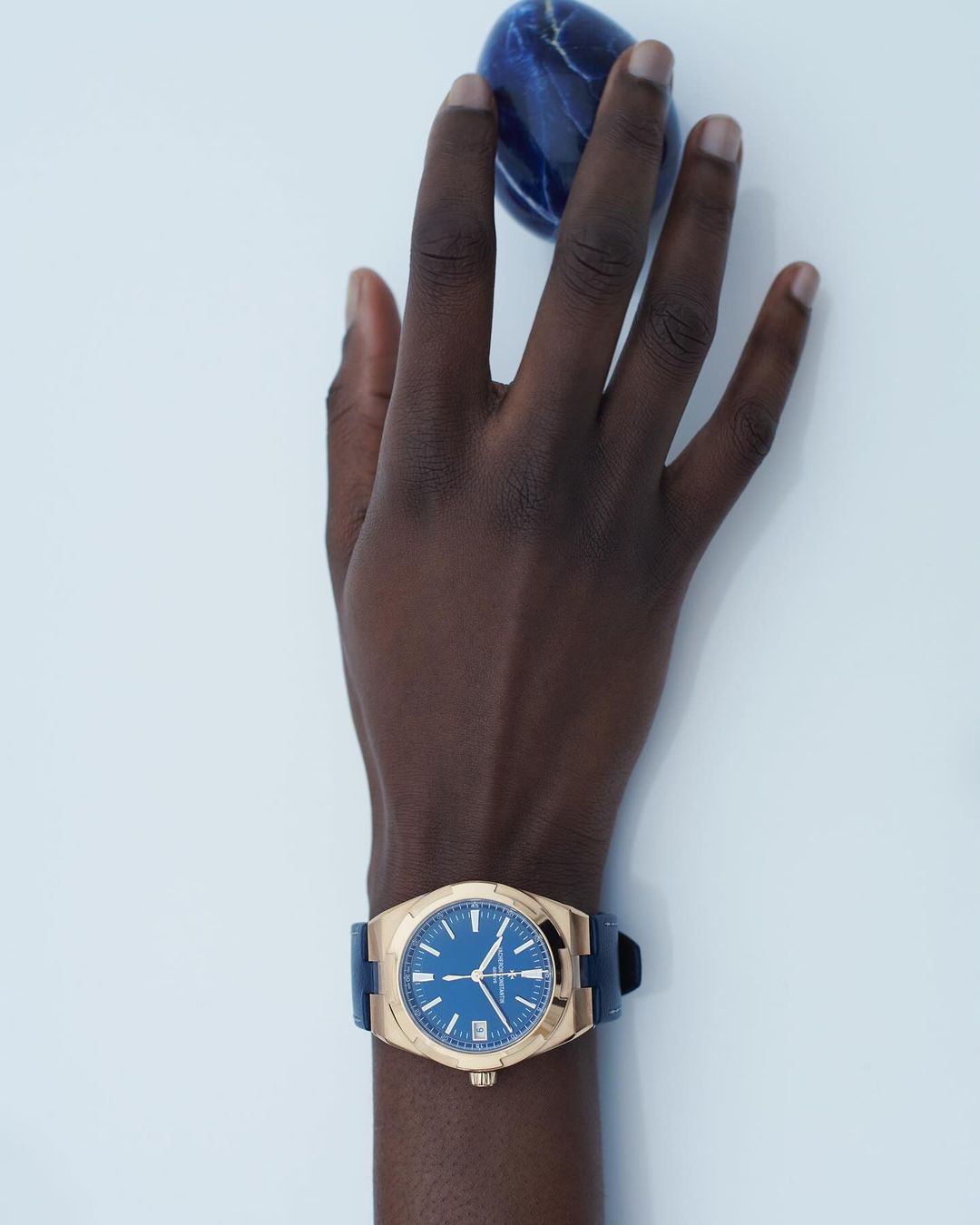 Hand with dark skin wearing a gold watch with blue dial. The hand is posed against a white backdrop, holding a dark blue stone.