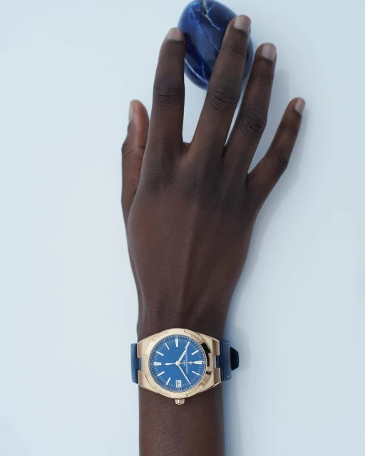 Hand with dark skin wearing a gold watch with blue dial. The hand is posed against a white backdrop, holding a dark blue stone.