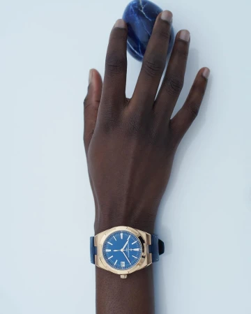 Hand with dark skin wearing a gold watch with blue dial. The hand is posed against a white backdrop, holding a dark blue stone.