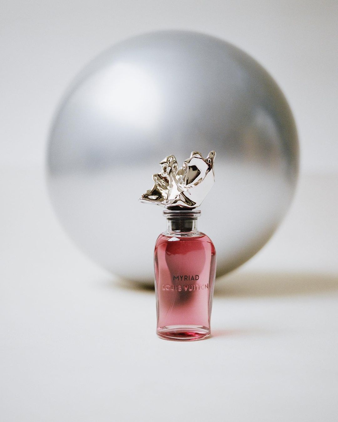 Louis Vuitton perfume bottle, Myriad, with silver decorative stopper against blurred silver sphere, product shot, beauty, indoors.