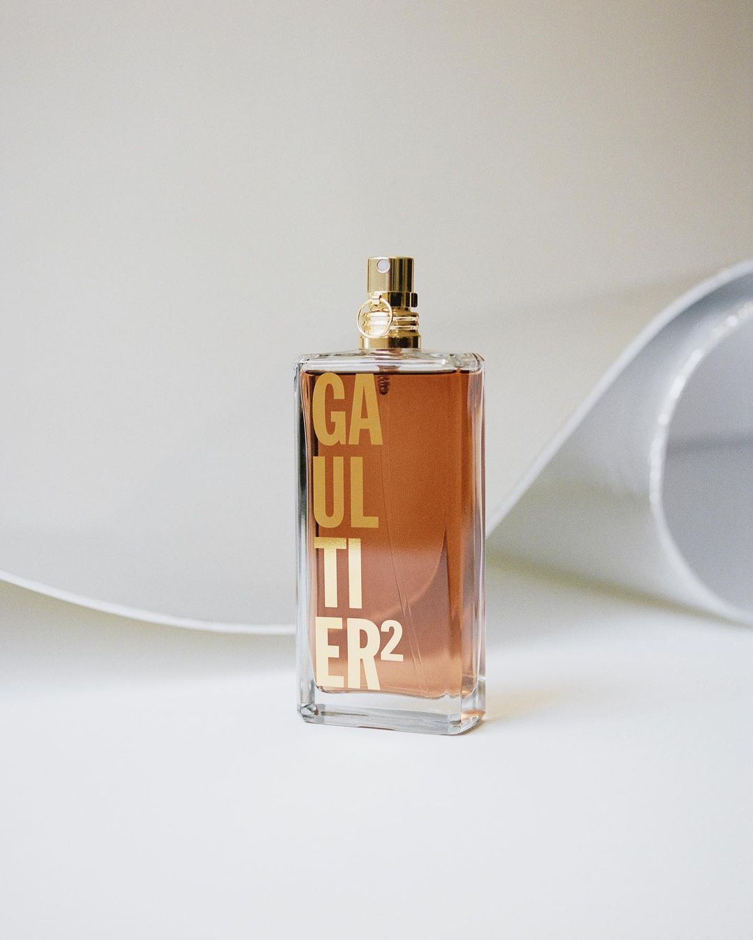 A clear glass perfume bottle with gold lettering and hardware stands on a white surface with a curved white backdrop in a studio setting.