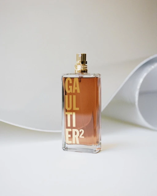 A clear glass perfume bottle with gold lettering and hardware stands on a white surface with a curved white backdrop in a studio setting.